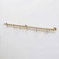 Angled full view of a long wall-mounted unlacquered solid brass kitchen rail with six S-hooks against a plain white background.