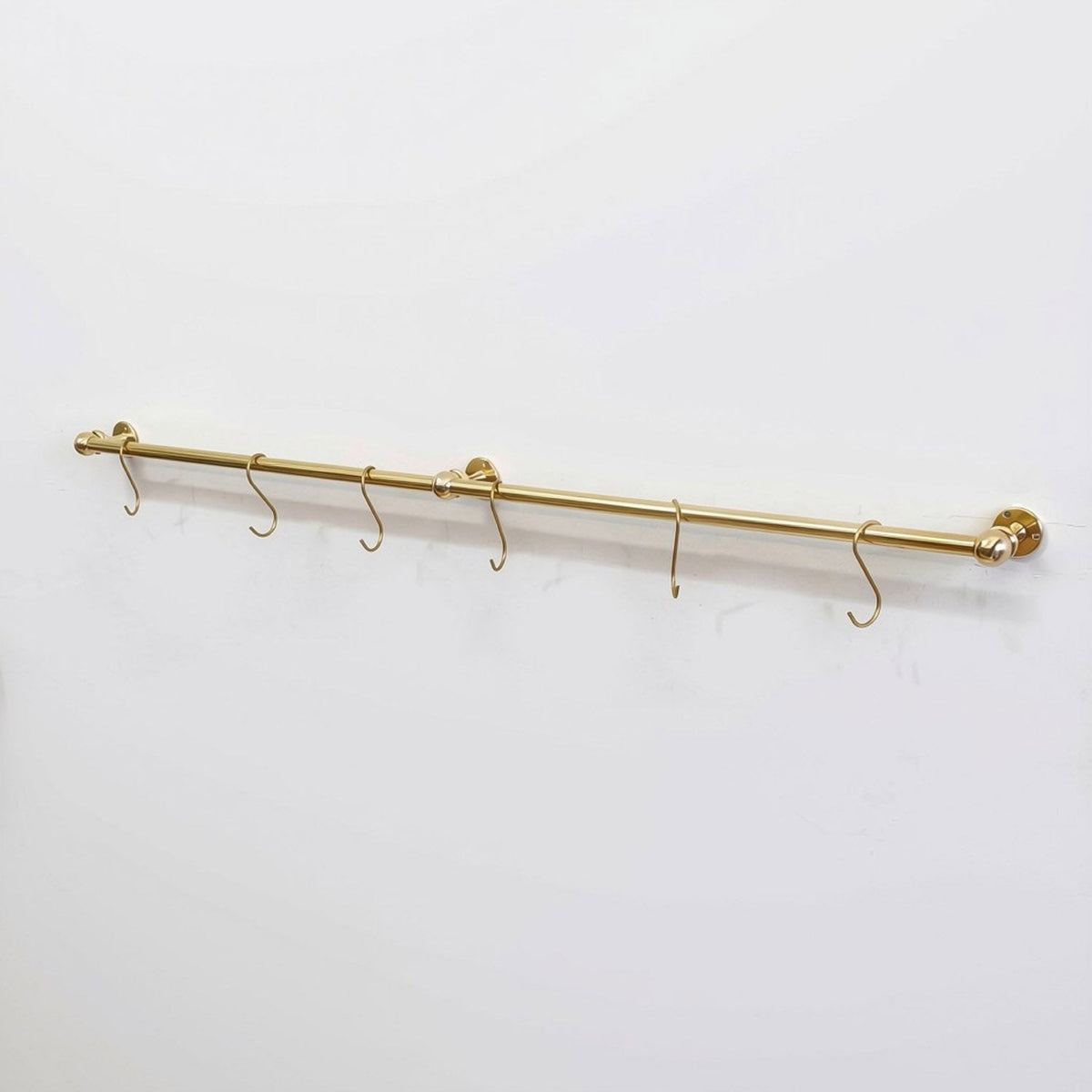Angled full view of a long wall-mounted unlacquered solid brass kitchen rail with six S-hooks against a plain white background.
