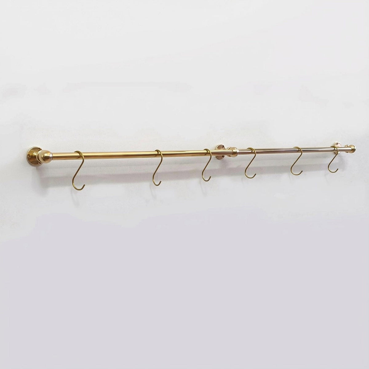 Full view of a long solid unlacquered brass kitchen hanging rail mounted on a plain white wall with six brass S-hooks.