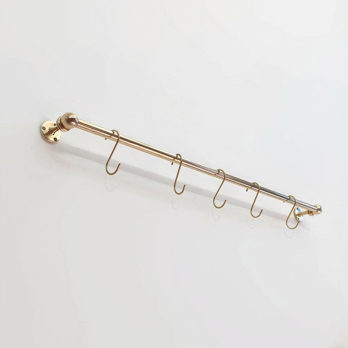 Perspective view of a short solid unlacquered brass kitchen hanging rail mounted on a plain white surface with five matching S-hooks.