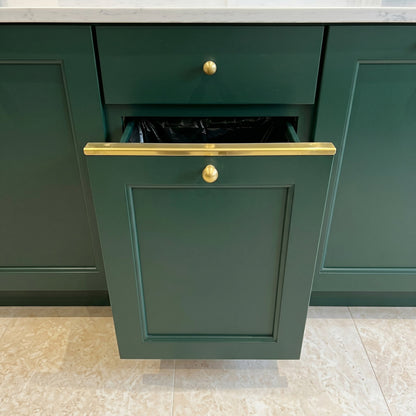 Solid brass drawer topper L-shaped edge guard installed on dark forest green kitchen cabinets with brass knobs and pull-out trash bin.