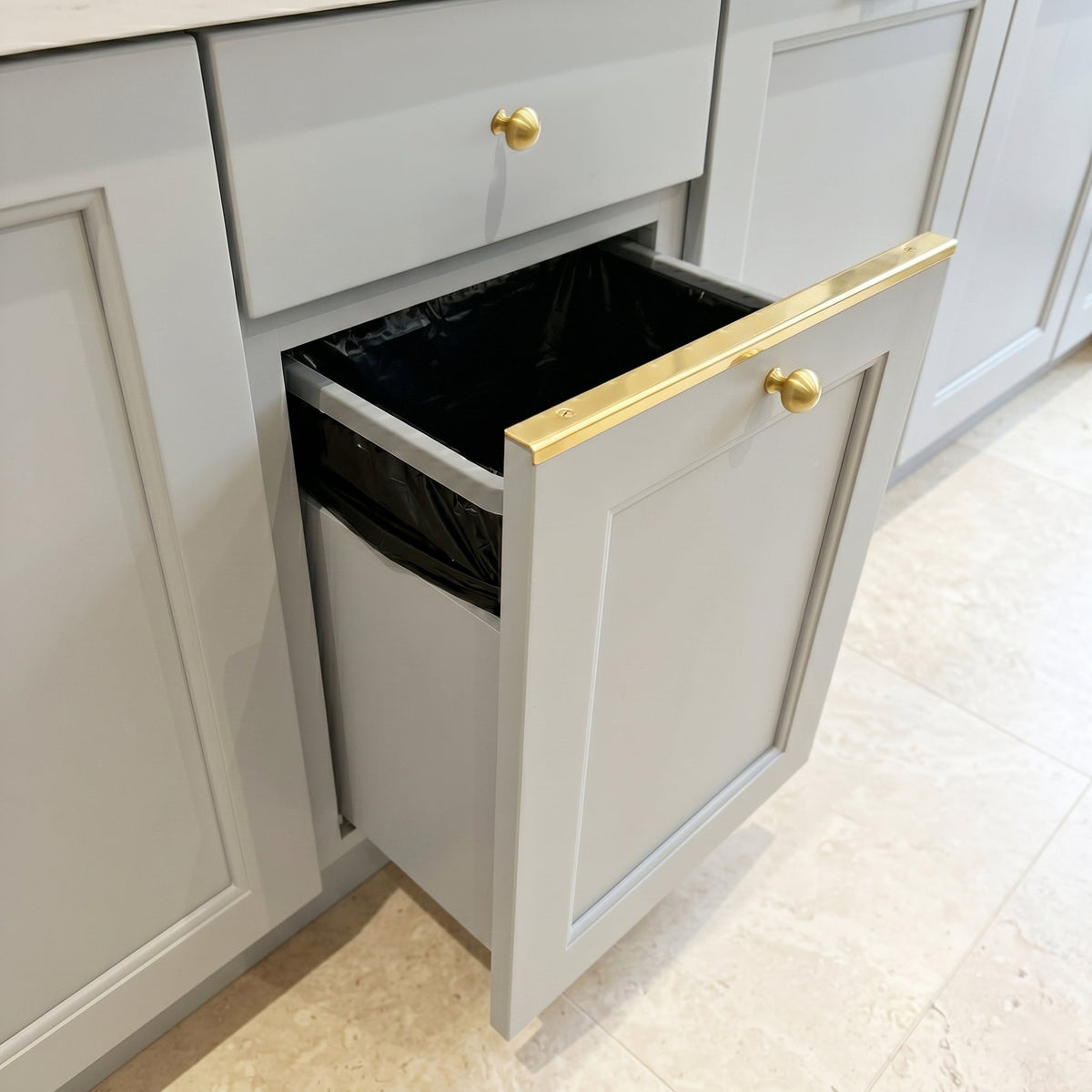 Pull-out trash cabinet with solid brass drawer topper L-profile protector and brass knob installed on light grey kitchen cabinets.