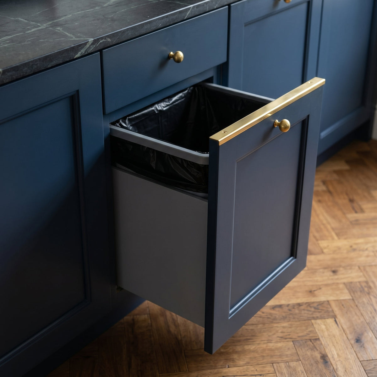 Pull-out kitchen trash cabinet featuring a solid brass edge protector topper and brass knob hardware on navy blue shaker cabinets.