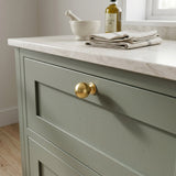 Unlacquered satin brass solid ball drawer knob installed on a sage green shaker cabinet vanity with a white marble countertop.