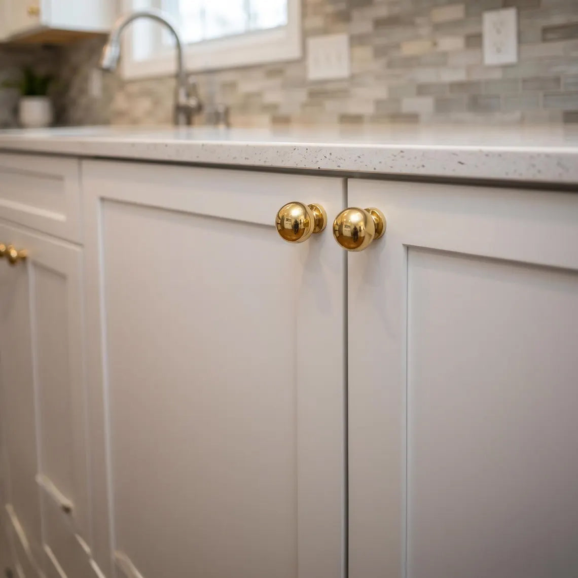 Unlacquered polished solid brass spherical ball knobs installed on classic white shaker kitchen cabinets with quartz countertop.