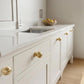 Unlacquered solid brass ball cabinet knobs mounted on off-white shaker drawers along a marble kitchen countertop.