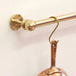 Close-up of a solid unlacquered brass kitchen rail end flange and ball finial on a white wall with an S-hook holding a copper pan.