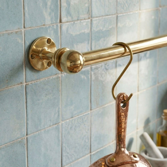 Close-up of a solid unlacquered brass kitchen rail mounted on light blue zellige tiles with a brass S-hook holding a hammered copper pan.