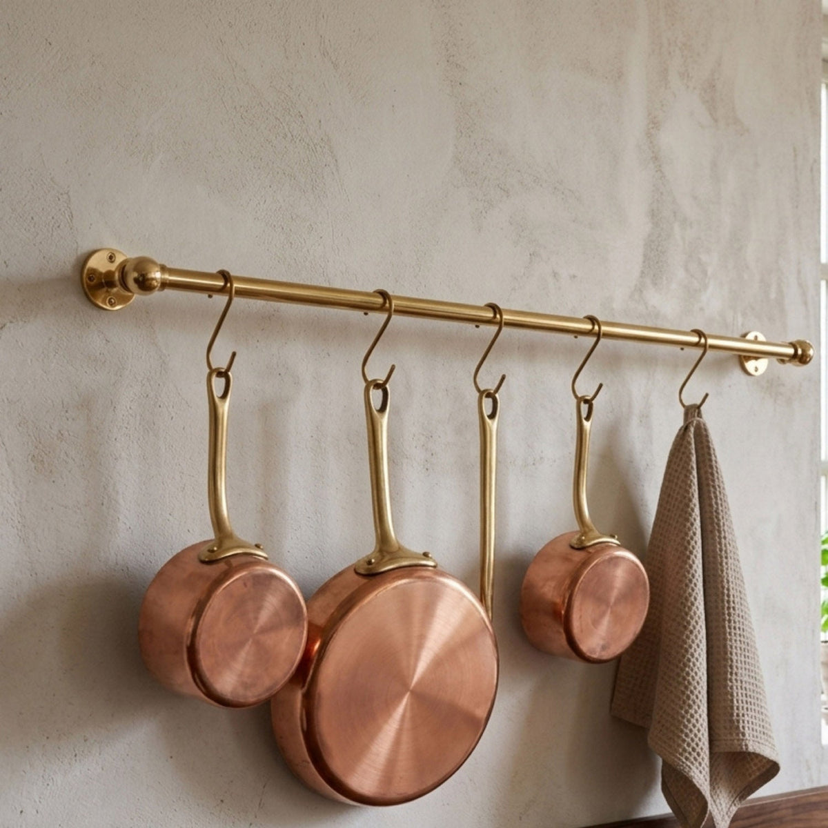 Unlacquered solid brass kitchen hanging rail mounted on a textured wall, displaying three handmade copper saucepans with brass handles and a beige waffle weave towel attached via S-hooks.