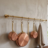 Unlacquered solid brass kitchen hanging rail mounted on a textured wall, displaying three handmade copper saucepans with brass handles and a beige waffle weave towel attached via S-hooks.
