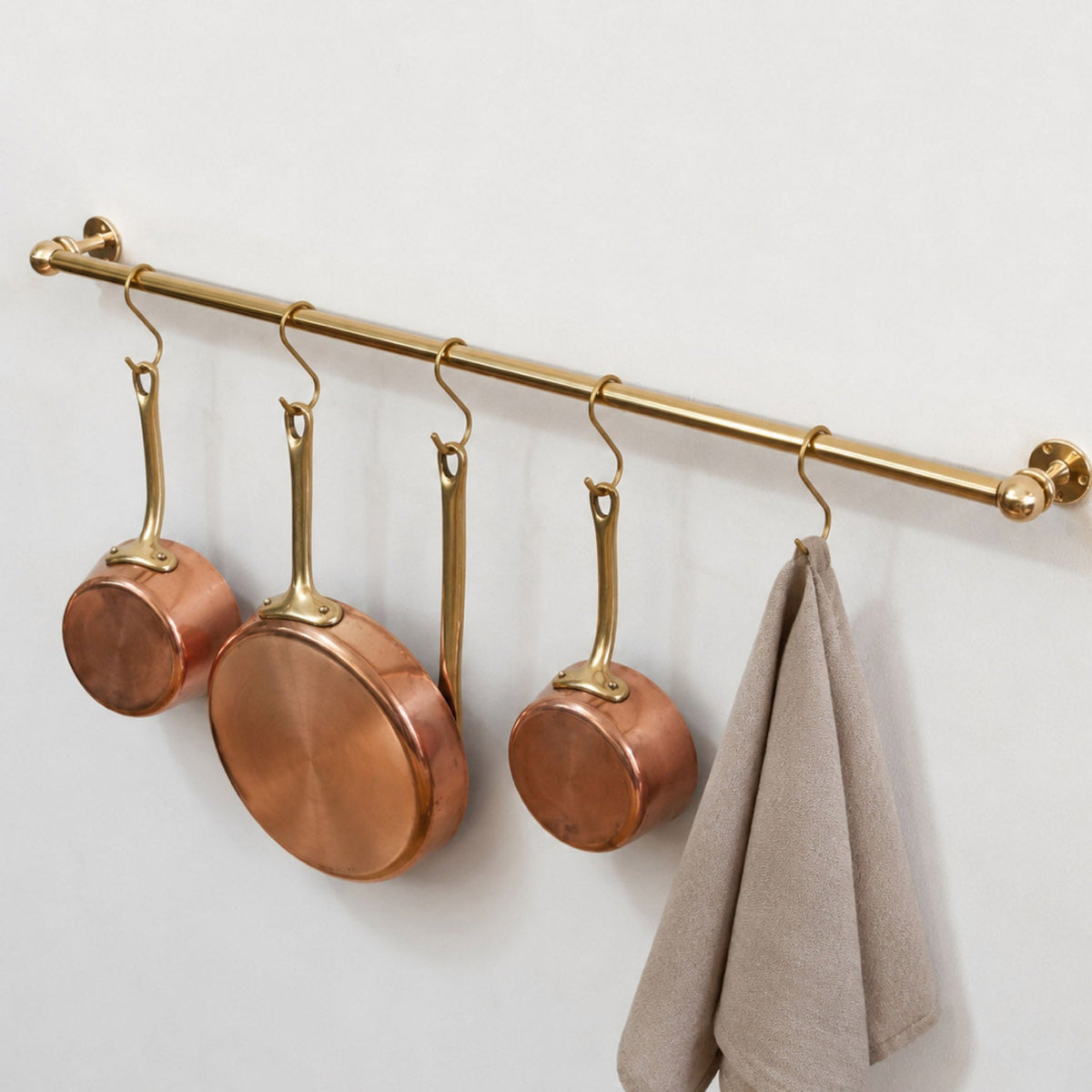 Solid unlacquered brass kitchen hanging rail mounted on a plain white wall holding copper pots, pans, and a linen towel with brass S-hooks.